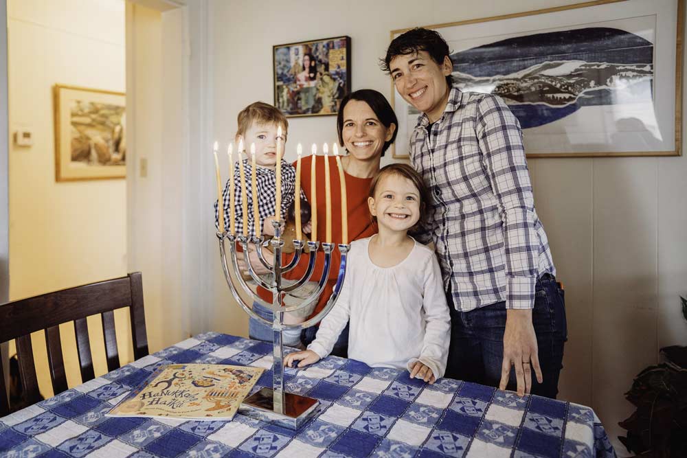 Two small children pose with their moms and a lit Hanukkah menorah.