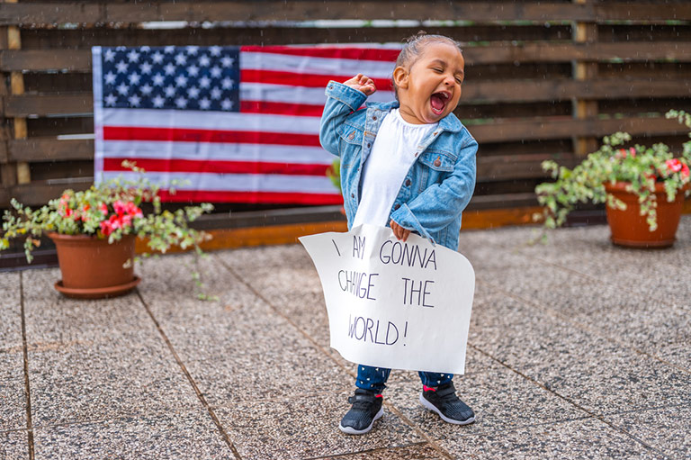 A child standing in front of the US flag with their arm raised victoriously over their head while holding a sign that says 'I'm going to change the world'