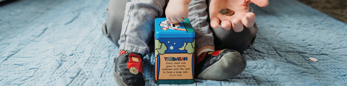 a tzedakah box rests on the floor and a toddler's hand drops a coin inside