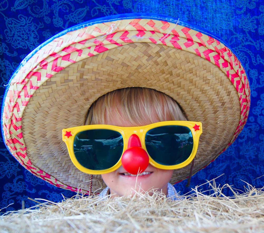A child in front of a bale of straw wearing a large western hat, large glasses and a large clown nose.