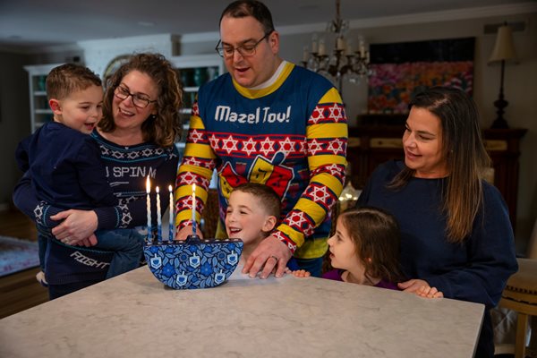 A family and friends getting together for Hanukkah. Two of the parents wear ugly holiday sweaters. The children smile and laugh. A menorah is lit.