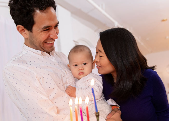 A mother and father holding their baby in front of a menorah