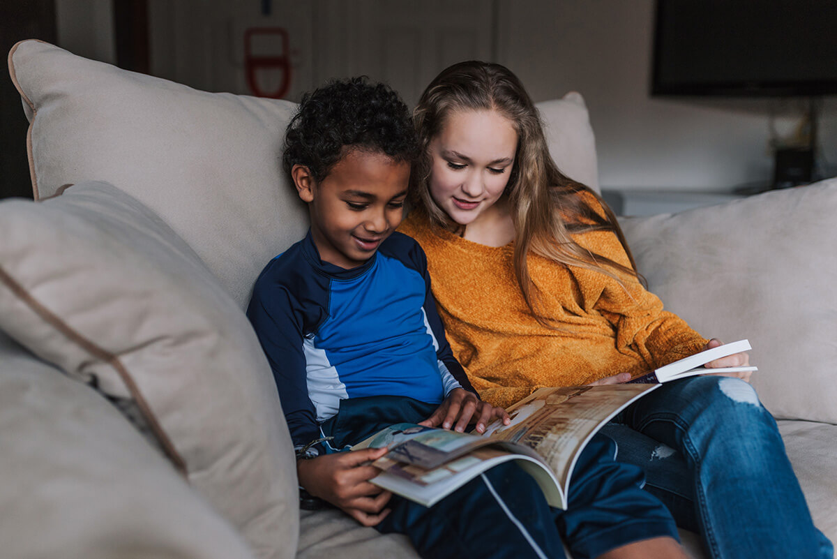 A girl and boy sit on a white couch and read an illustrated book together.
