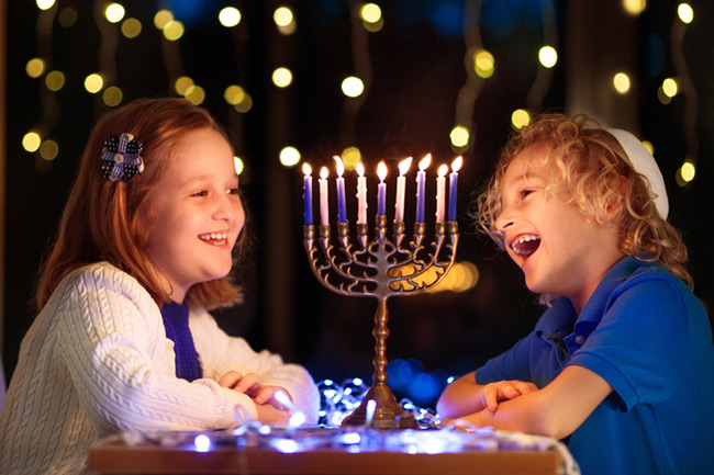 Two children sitting at a table looking at a lit menorah and smiling