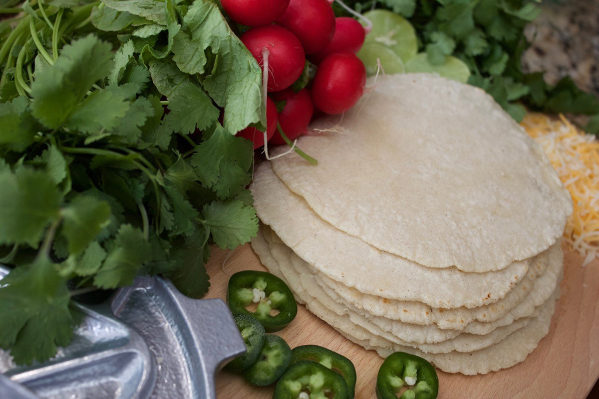 Corn tortillas on table surrounded by greens, peppers, and tomatoes
