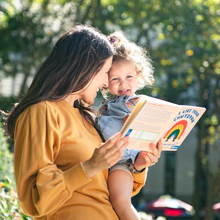 A woman reads a book with her child outdoors in the sun