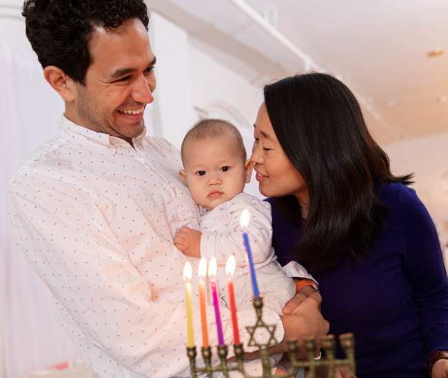 Parents holding their baby in front of a partially lit menorah